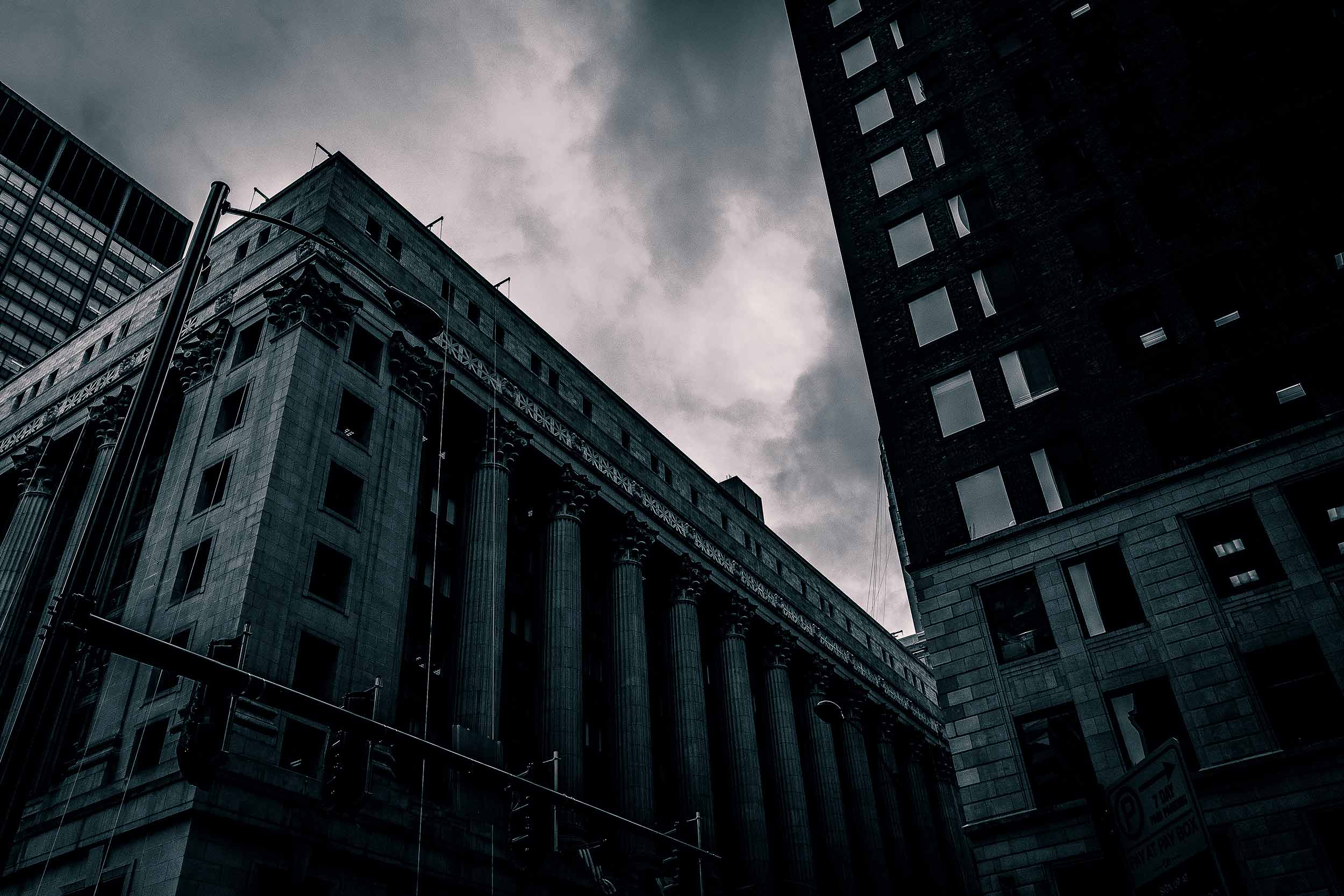 black and white image of an angle looking up at the corner of a solid building in a big city with an ominous cloudy sky in the background